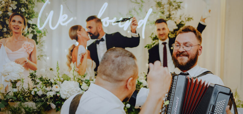 Gitarzysta i akordeonista szaleją i grają na kolanach biesiadny utwór przy stole pary młodej na weselu w Hotelu Nowodwory.