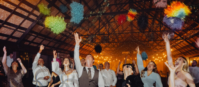 Goście weselni podrzucają do góry kolorowe peruki które dostali od DJ'a podczas zabawy. Wszyscy mają wyciągnięte ręce do góry a peruki lecą pod sufitem. DJ też tam jest.