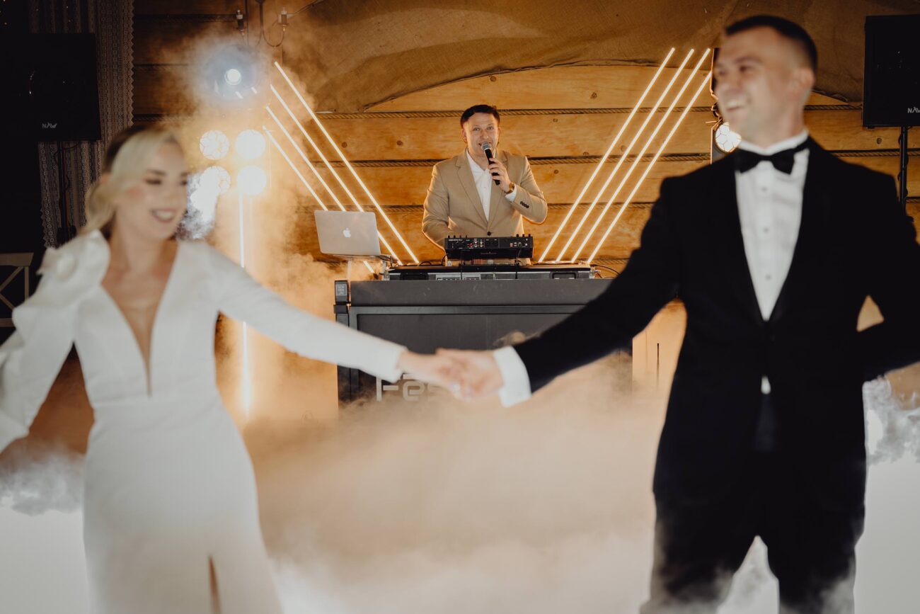 Zdjęcie DJ-a Marcina Brzozowskiego uchwycone podczas 1 tańca młodej pary. Marcin znalazł się idealnie między nimi. Oni trzymają się za ręce a Marcin patrzy się w obiektyw mówić coś do mikrofonu.