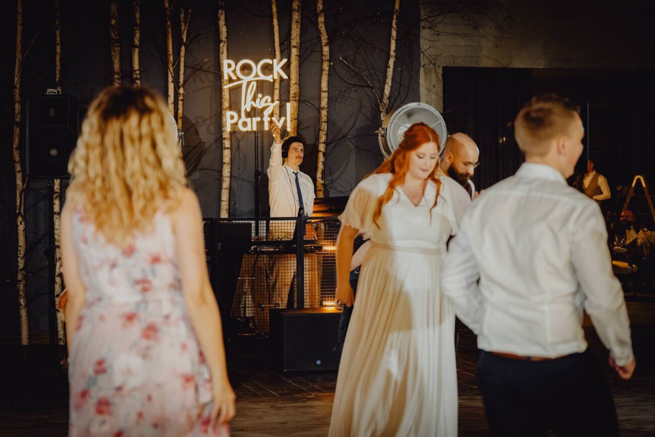 Wodzirej na weselu macha do ludzi zza konsolety grając muzykę.