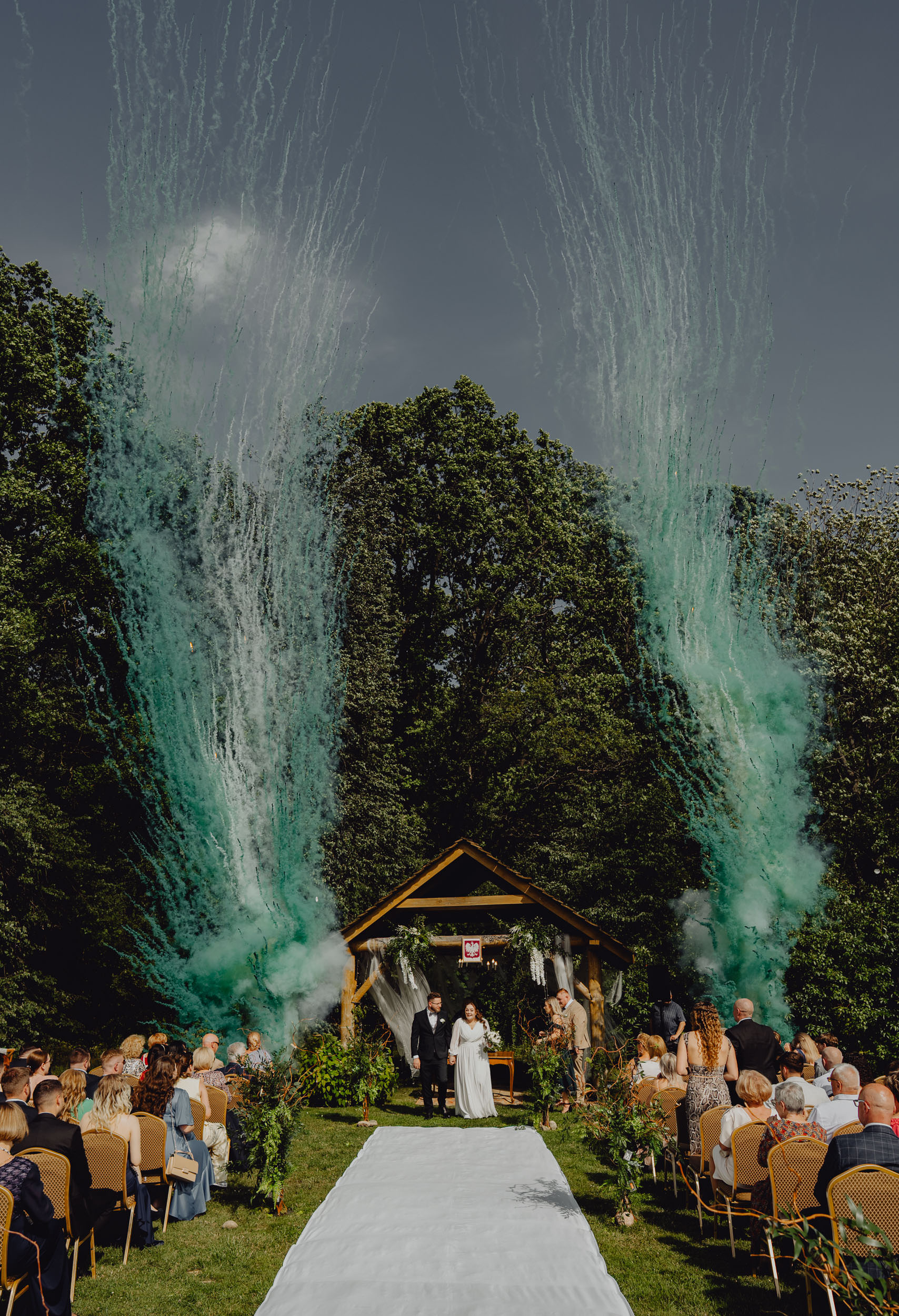 Para Młoda idzie po białym dywanie a za nią wybuchają dwie ogromne fontanny dymne. Ceremonia ślubna się zakończyła, goście biją brawo.