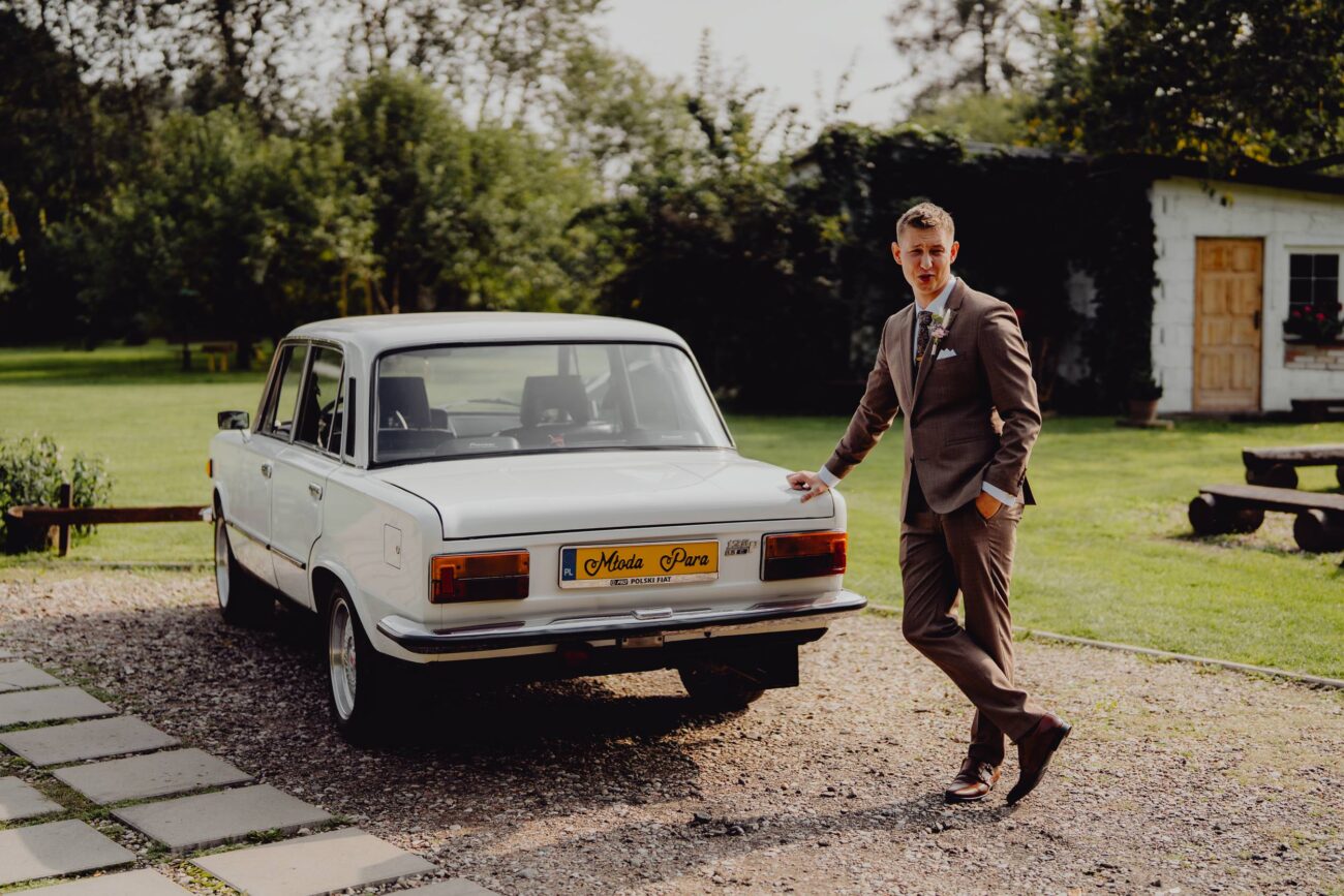 Pan Młody opiera się o białe auto do ślubu fiat 125p