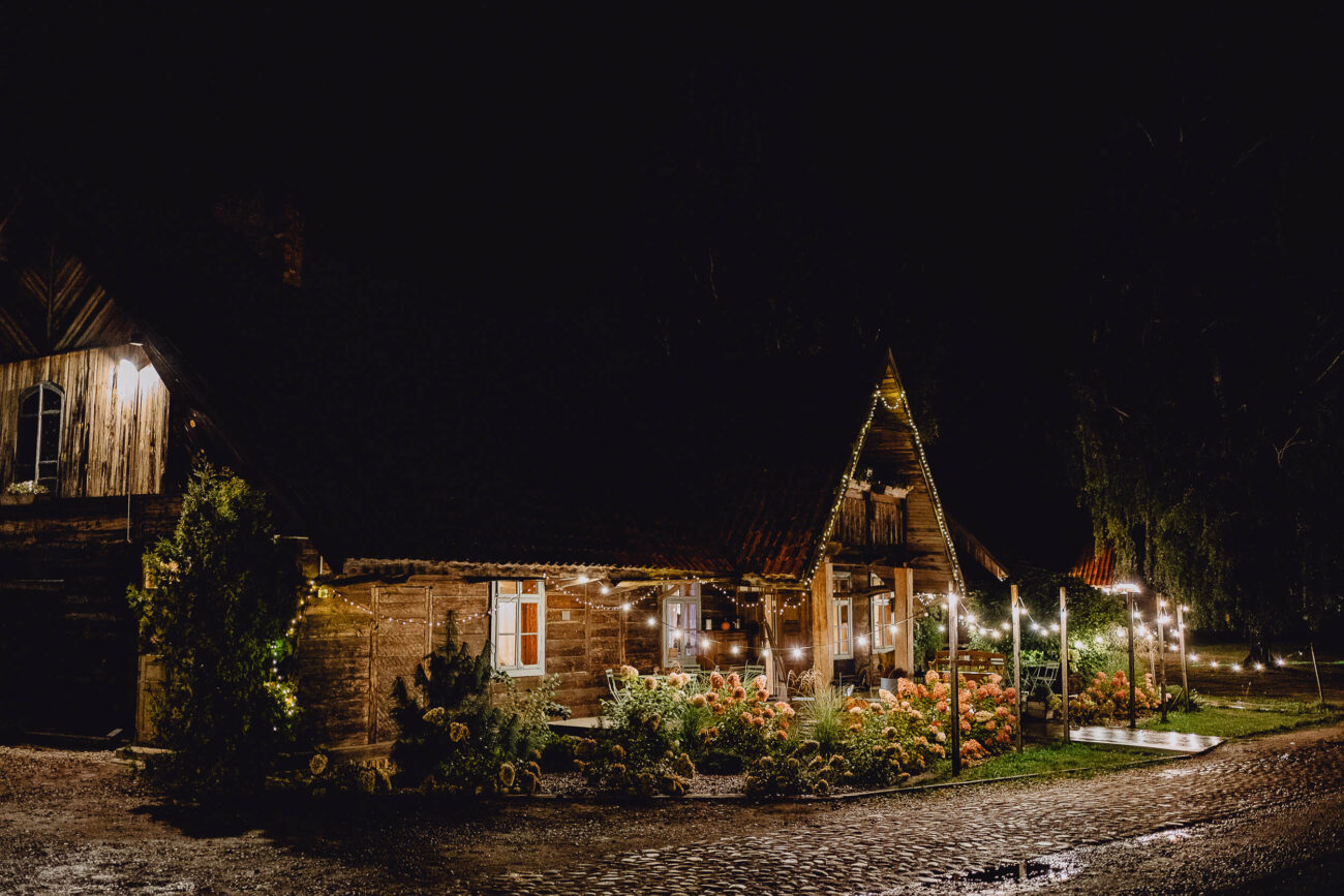 Stodoła Weselna Dworek Rustykalny sfotografowany w nocy. Dookoła piękne lampki i zieleń.