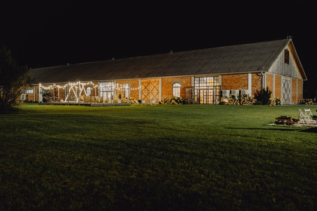 Zdjęcie Ekskluzywnej Stodoły Weselnej Majątku Howieny od strony ogrodu. Widać piękny ogród, trawnik, drewniną oranżerię i ozdobne oświetlenie. Zdjęcie wykonane w nocy.