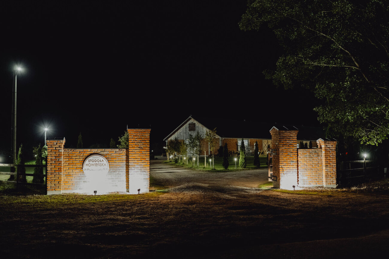 Brama wjazdowa do Stodoły Howieńskiej wykonana ze starej cegły. Widac podświetlane logo Majątku Howieny oraz zieleń i piękne ozdobne ośiwetlenie.